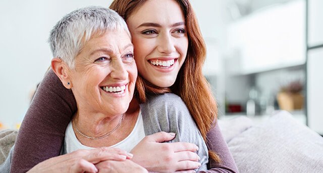 Pflege-Ratgeber Junge Frau umarmt ältere Damen von hinten beide lächeln